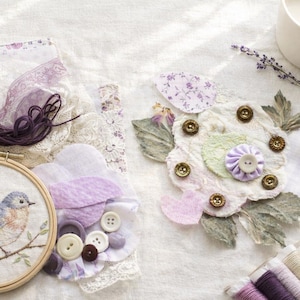 May include: An embroidery hoop with a bluebird design, lace, fabric flowers, buttons, and spools of thread. The color palette includes shades of purple, white, and green, suggesting a craft or DIY theme.