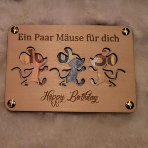 Puede incluir: Una tarjeta de cumpleaños de madera con tres recortes de ratones, cada uno lleno de papel de colores. La tarjeta dice "Ein Paar Mäuse für dich" y "Happy Birthday". Tornillos dorados aseguran las esquinas.