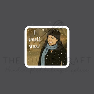 May include: Square sticker featuring a smiling person wearing a black coat, blue scarf, and hat, with the text "I smell snow." The background has a snowy effect. The sticker is a handcrafted baking supply.