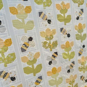Könnte beinhalten: Eine handgefertigte Steppdecke mit einem sich wiederholenden Muster aus gelben und weißen Blumen und Bienen. Die Blumen haben gelbe Blütenblätter und grüne Stiele und Blätter. Die Bienen sind schwarz, weiß und gelb. Die Steppdecke ist vor einem weißen Hintergrund.