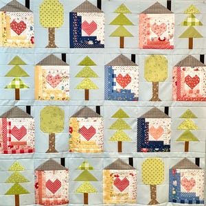 Peut inclure: Une courtepointe faite à la main avec un motif répété de maisons et d'arbres. Chaque maison présente un cœur et un toit gris. Les arbres sont verts et bruns, sur un fond bleu clair. Fabriqué à partir de divers tissus à motifs.