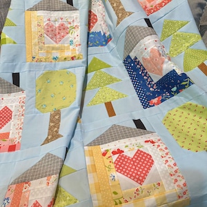 May include: Handmade quilt featuring a charming house and tree design. The quilt showcases a light blue background with appliqued houses, trees, and hearts in various fabrics. The houses have gray roofs and the hearts are red.