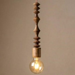 Puede incluir: Una lámpara colgante de madera con un cuerpo decorativo de madera tallada y una bombilla redonda. La luz cuelga de un cable rojo. La lámpara es de color madera natural y cálido.