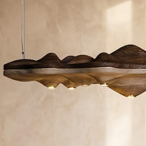 May include: A modern, sculptural chandelier hangs above a wooden dining table. The light fixture has a dark brown, mountain-like top and a lighter, textured underside with integrated lights. The table is set with plates and napkins, suggesting a dining setting.