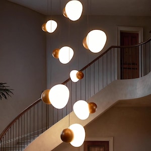 May include: Modern chandelier with multiple glowing, oval-shaped lights suspended from a wooden ceiling mount. The lights are white and gold, creating a warm ambiance. The fixture hangs in a grand entryway with a curved staircase.