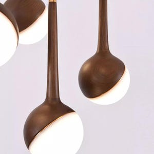 Puede incluir: Primer plano de lámparas colgantes modernas. Cada lámpara presenta un tallo de madera marrón oscuro y una pantalla esférica, dividida diagonalmente con una sección blanca y brillante. Una fina banda dorada acentúa el borde de la esfera. Las luces cuelgan sobre un fondo blanco.