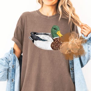 May include: Brown t-shirt featuring a patchwork duck design. The duck has a green plaid head, a brown floral patterned body, and wings with various fabric patterns. The shirt has rolled-up sleeves and a relaxed fit. The text "KS DESIGN STUDIO" is visible.