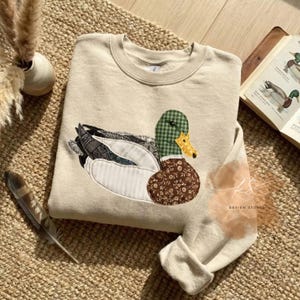 Peut inclure: Un sweat-shirt beige avec un motif de canard appliqué. Le canard a une tête à carreaux verts, un bec jaune et un corps à motifs floraux marron. Les ailes sont un mélange de tissus gris, blancs et verts. Le motif est sur un sweat-shirt de couleur neutre.