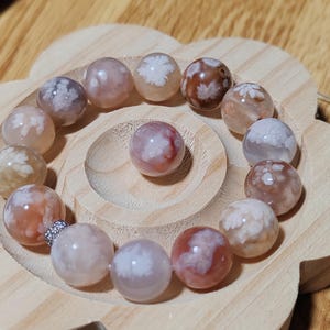 Puede incluir: Un brazalete hecho de cuentas redondas de ágata blanca, rosa y marrón con un cierre plateado. Las cuentas tienen un patrón único, nublado y veteado.