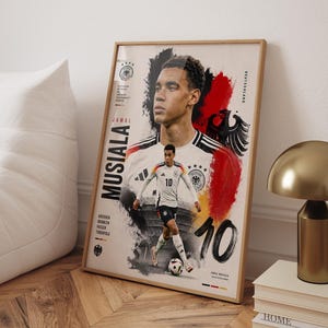 May include: A framed poster featuring a portrait and action shot of soccer player Jamal Musiala in a Germany jersey. The design includes the text 'JAMAL MUSIALA', the number '10', the German eagle emblem, and black, red, and gold paint splatters.