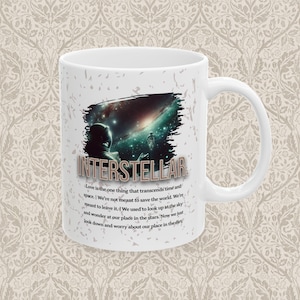 May include: White ceramic mug featuring the word "INTERSTELLAR" with a space-themed graphic. The design includes a person in a spacesuit and a nebula. Text on the mug reads, "Love is the one thing that transcends time and space."