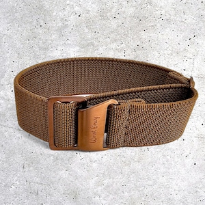 Peut inclure: Un bracelet de montre marron avec une boucle de couleur bronze. Le bracelet est fait d'un matériau tissé. La boucle porte l'inscription "Wrist Envy". Le bracelet de montre est présenté sur un fond gris.