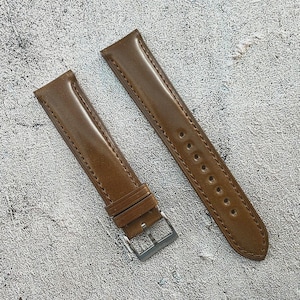 May include: Two brown leather watch straps with a shiny finish. One strap has a silver buckle and a loop to hold the strap in place. The other strap has multiple holes for adjustment. The straps are lying on a textured, light gray surface.