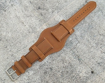 May include: A brown leather watch strap with a unique design. The strap features a wide central section with two smaller straps and a silver buckle. The leather has visible stitching and a textured surface.