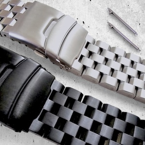 Könnte beinhalten: Nahaufnahme von zwei Edelstahl-Uhrenarmbändern, eines silberfarben und eines schwarz. Das silberfarbene Band ist im Fokus und zeigt einen Verschluss und ein Design mit gewebten Gliedern. Zwei silberne Stifte sind sichtbar. Das schwarze Band befindet sich darunter, ebenfalls mit einem gewebten Gliederdesign.