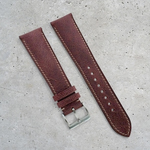 May include: Burgundy leather watch strap, two-piece design. The strap has cream stitching and a silver buckle. The leather has a textured finish. The strap is designed for a wristwatch.