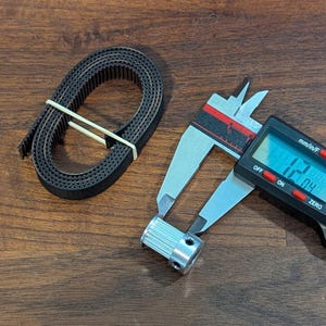 May include: A black timing belt coiled and secured with a rubber band, next to a digital caliper measuring a silver pulley. The caliper displays a measurement of 12.04 mm. The items are on a wooden surface.