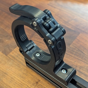 May include: Black plastic clamp with a locking mechanism and a threaded ring. The clamp is attached to a black metal rail.