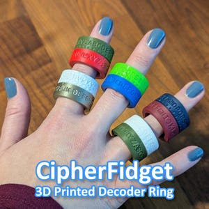 Puede incluir: Una mano que lleva varios anillos decodificadores impresos en 3D de colores. Los anillos son de varios colores, incluyendo rojo, verde, azul, blanco y marrón, cada uno con un alfabeto grabado. El texto "CipherFidget 3D Printed Decoder Ring" está en la parte inferior.