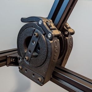 May include: Close-up of a black 3D printed camera slider with two adjustable joints. The joints have a locking mechanism with a lever and a circular knob. The slider is made of aluminum extrusion.