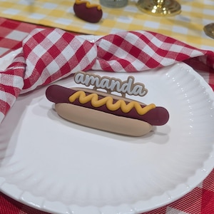 May include: A decorative hot dog on a white plate with a red and white checkered napkin. The hot dog has a brown bun, a red sausage, and yellow mustard. The name "amanda" is above the hot dog.