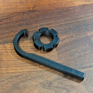 May include: Black, textured 3D-printed tool set. The set includes a curved hook-shaped tool and a circular, gear-like component. The tools are displayed on a wooden surface.