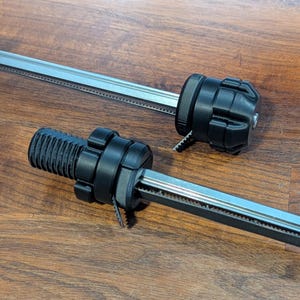 Op de afbeelding: Twee zwarte en zilveren lineaire bewegingscomponenten worden getoond op een houten oppervlak. Elk heeft een zwart, cilindrisch uiteinde met een geribbelde textuur en een zilveren, getande rail. Deze componenten zijn ontworpen voor precieze lineaire beweging.