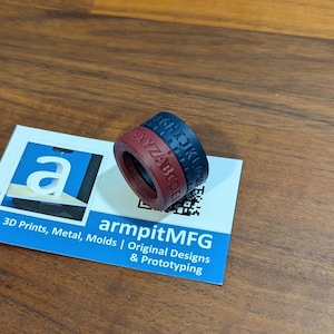 Pode incluir: Um anel impresso em 3D com duas faixas, uma azul escuro e outra bord&ocirc;, cada uma com letras gravadas. O anel est&aacute; sobre um cart&atilde;o de visita com o texto "armpitMFG" e "3D Prints, Metal, Molds | Original Designs & Prototyping".