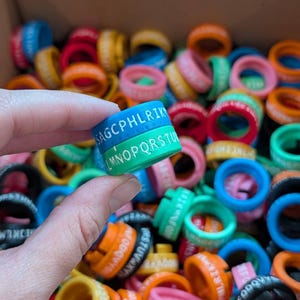 May include: A collection of colorful silicone rings, each with a band of white letters. One ring is held up, displaying the letters "SAGCPHLRIK" and "LMNOPQRSTU" in white. The rings are in various colors, including blue, green, pink, and orange.