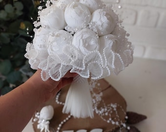 May include: A round bouquet of white artificial peony flowers accented with small white pearls and delicate white tulle ruffles. The bouquet features a white fabric-wrapped handle and is decorated with strands of white pearls throughout the floral arrangement.