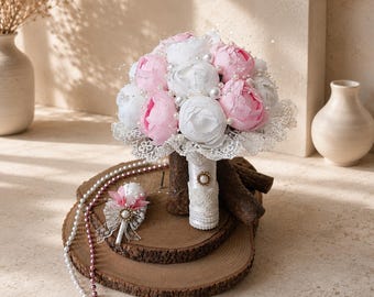 May include: A bouquet of artificial pink and white peonies accented with small white pearls and delicate lace trim. The handle is wrapped in white satin ribbon with a pearl embellishment. A matching boutonniere features a single peony and lace.