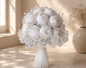 May include: A round bouquet of white artificial peonies accented with small white pearls on thin stems. The base of the bouquet is wrapped in white fabric with a ruffled lace trim and a long, straight white tassel handle adorned with pearls.