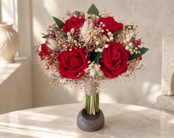 May include: A floral bouquet featuring deep red roses, dried baby's breath, and small white bunny tail grass. The stems are bundled together with natural twine and held upright in a dark, rounded, smooth-surfaced base, creating a compact arrangement.