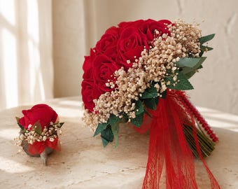 May include: A bouquet of deep red roses accented with small white baby's breath flowers and green leaves, tied with a red lace ribbon. Beside it sits a matching boutonniere featuring a single red rose, baby's breath, and a small red ribbon.