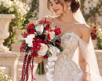 Op de afbeelding: Een bruidsboeket met dieprode rozen, witte pioenrozen en donkerrode anthuriums, geaccentueerd met lange, hangende rode amarantstelen en groen blad. Vastgehouden door een persoon in een witte, strapless kanten trouwjurk.