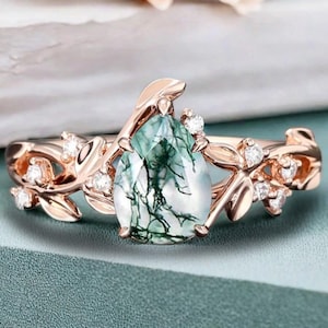 May include: Rose gold ring featuring a pear-shaped moss agate gemstone with green and white veining. The ring has a floral design with small, clear accent stones and leaf-shaped details. The ring is set against a teal background.