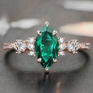 Puede incluir: Un anillo de oro rosa con una piedra central de esmeralda con corte marquesa. El anillo está adornado con pequeñas piedras blancas redondas a cada lado de la esmeralda. La banda también está detallada con pequeñas piedras preciosas. Una hermosa joya.