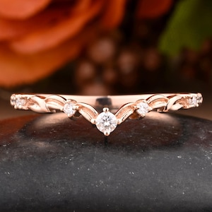 May include: Rose gold ring with a curved design. The ring features a central, round, clear gemstone and smaller gemstones along the band. The band has a delicate, leaf-like pattern. The ring is displayed on a dark, textured surface.