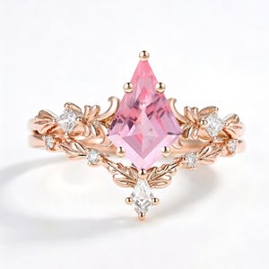 May include: A rose gold ring set featuring a large kite-shaped pink center stone held by prongs. The band is decorated with intricate leaf-like metalwork and small clear square and kite-shaped accent stones embedded along the ornate metal frame.