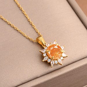 Puede incluir: Collar dorado con un colgante. El colgante tiene una piedra preciosa naranja central, rodeada de piedras más pequeñas y claras, formando un diseño de sol. La fina cadena y el colgante se muestran sobre una superficie de tono neutro. Joyería.