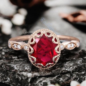 May include: A rose gold engagement ring featuring a large oval ruby gemstone, surrounded by a delicate filigree setting and smaller diamonds. The ring is displayed on a dark grey textured surface.