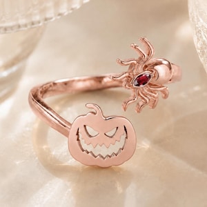 May include: A rose gold open ring featuring a carved pumpkin jack-o'-lantern on one end and a detailed spider with a small, marquise-cut red gemstone set into its body on the other end. The metal has a polished, reflective finish throughout.