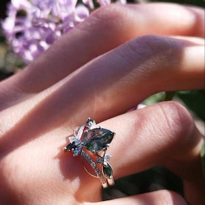 May include: A silver ring with a large, teardrop-shaped green and brown gemstone. The ring has a floral design with smaller gemstones and is worn on a finger. The background includes blurred purple flowers and greenery.