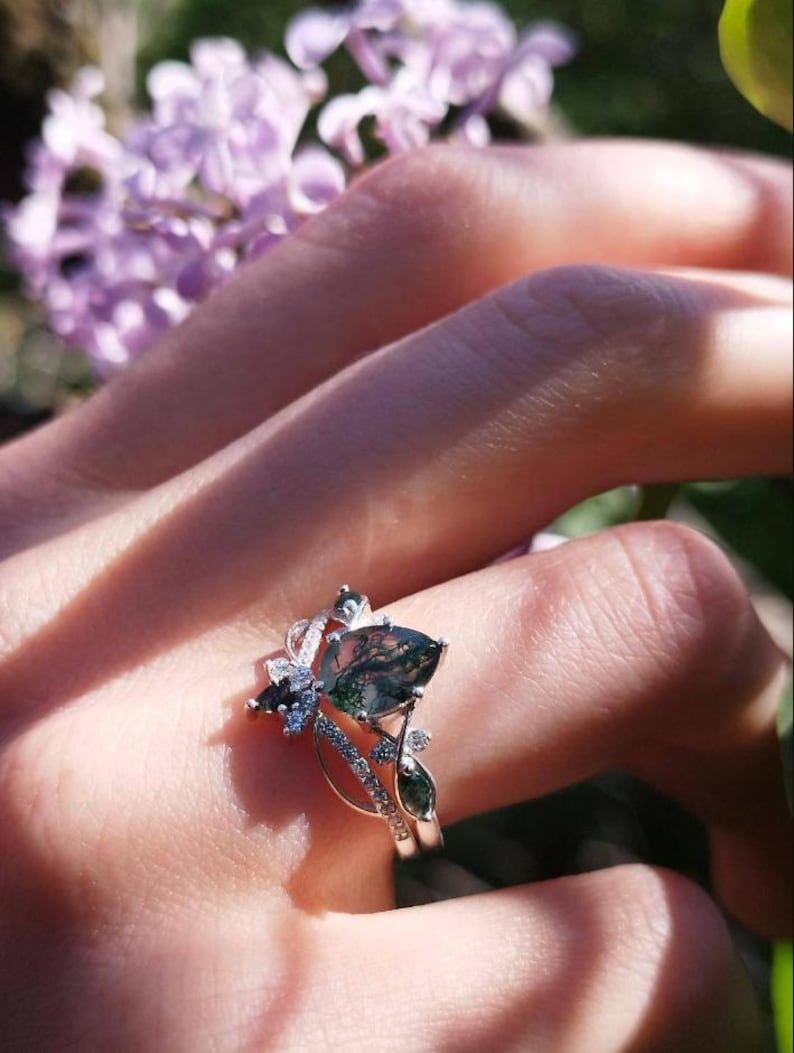 May include: A silver ring with a large, teardrop-shaped green and brown gemstone. The ring has a floral design with smaller gemstones and is worn on a finger. The background includes blurred purple flowers and greenery.
