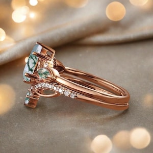 May include: Rose gold ring set featuring a large, oval-shaped moss agate stone with green and white patterns. The set includes a band with small, clear gemstones and a matching band. The rings are displayed on a neutral surface.