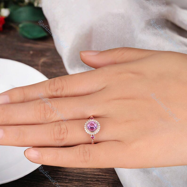 May include: A rose gold ring featuring a pink gemstone encircled by a halo of smaller diamonds. The band of the ring is adorned with two smaller pink gemstones.