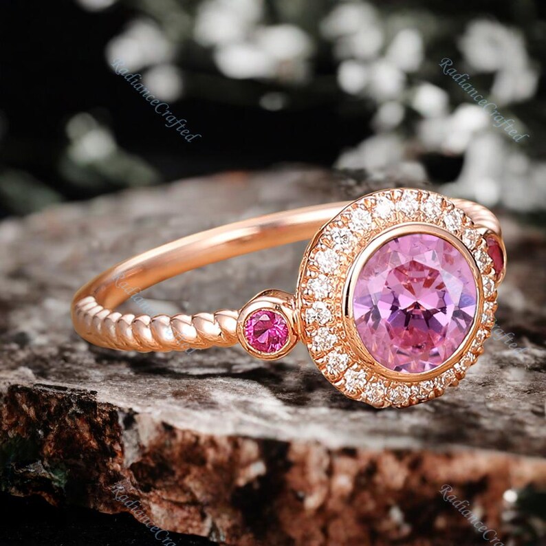 May include: A rose gold engagement ring featuring a pink gemstone encircled by a halo of smaller diamonds. The band is twisted and has two smaller pink gemstones on either side of the main stone.