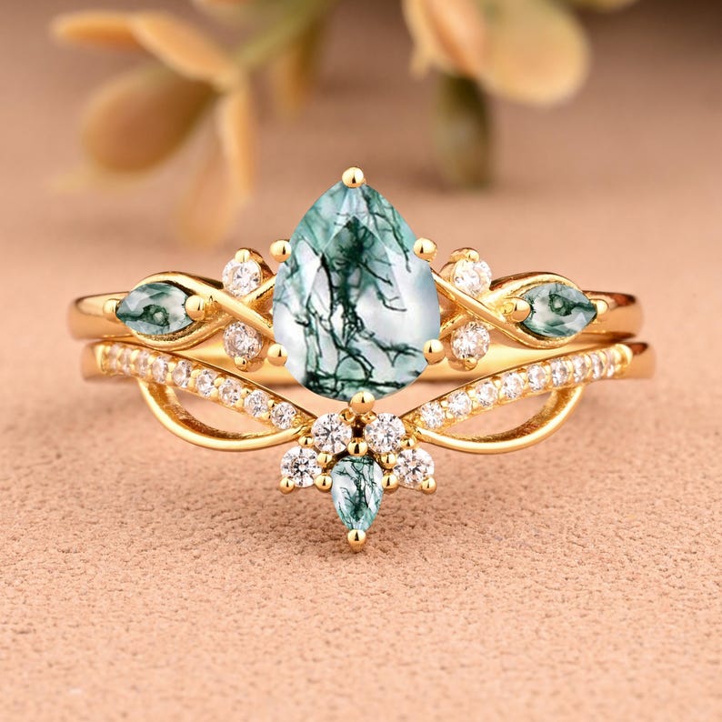 May include: A gold ring set featuring a pear-shaped, green moss agate gemstone as the centerpiece. The ring is accented with small, clear gemstones and additional green gemstones. The band has a delicate, ornate design.