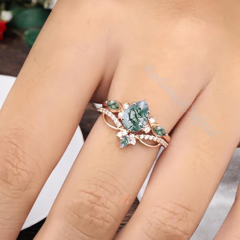 May include: Rose gold ring set featuring a pear-shaped moss agate gemstone with green inclusions. The engagement ring is accented with small white gemstones and two green gemstones. The wedding band has a delicate, curved design.