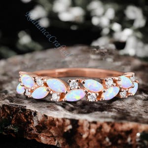 May include: A delicate rose gold band ring with a row of oval-shaped white opals and small round clear diamonds. The ring is set on a brown textured surface.